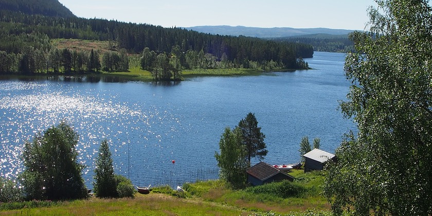 Två små båthus vid en strandkant, sjö och berg i bakgrunden.