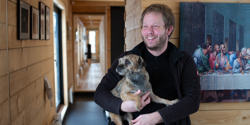 Peter med hunden berit i famnen, huset sträcker sig långt och smalt bakom honom.