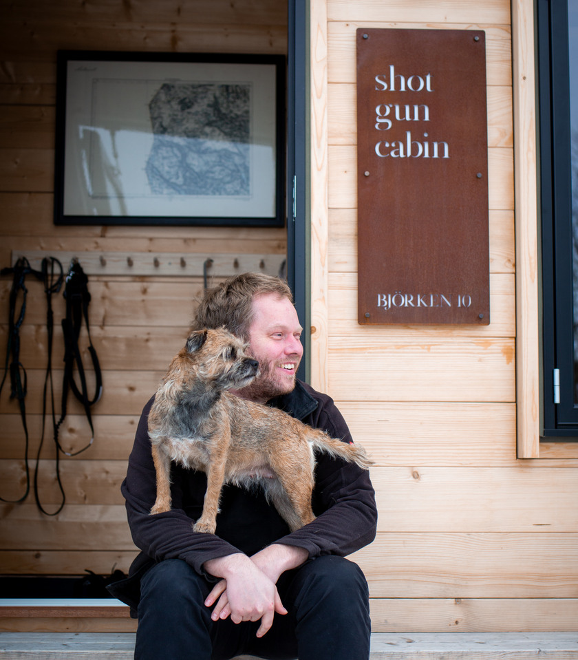 Peter sitter med sin hund berit i knät. Texten Shotgun cabin står på en rostig skylt bakom.