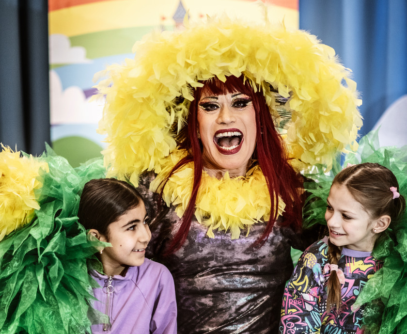 Sagostund med drag queen som har hatt av gula fjädrar och en glittrig klänning, två barn sitter bredvid.