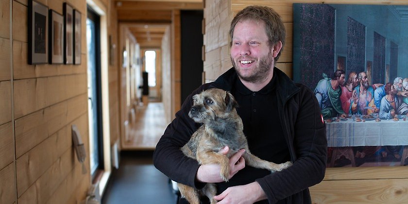Peter med hunden berit i famnen, huset sträcker sig långt och smalt bakom honom.